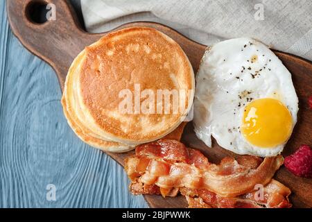 Brett mit leckeren Pfannkuchen, gebratenem Speck und Ei auf Holztisch Stockfoto