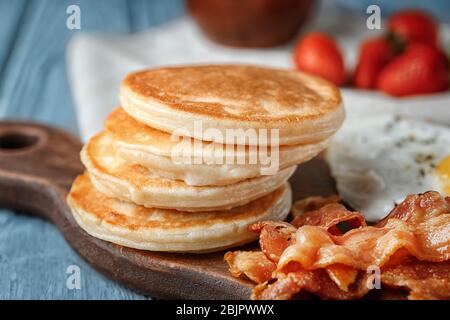 Brett mit leckeren Pfannkuchen, gebratenem Speck und Ei auf Holztisch Stockfoto