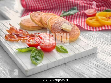 Brett mit leckeren Pfannkuchen, Tomaten und gebratenem Speck auf Holztisch Stockfoto