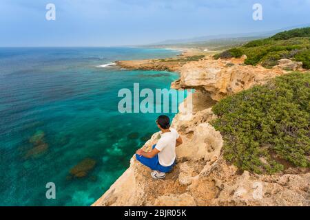 Junger Mann sitzt am Rande einer Klippe mit Blick auf das Meer am Lara Beach auf der Akamas Halbinsel, Zypern Stockfoto