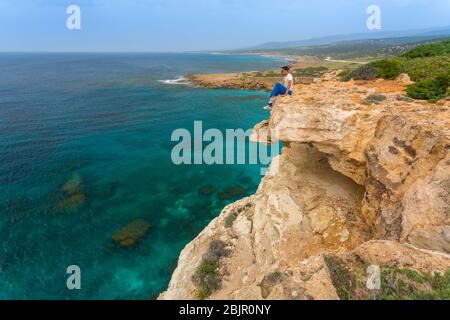 Junger Mann sitzt am Rande einer Klippe mit Blick auf das Meer am Lara Beach auf der Akamas Halbinsel, Zypern Stockfoto