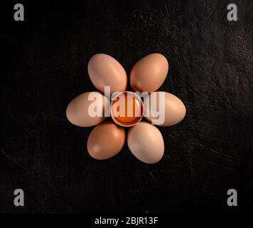 Flaches Hühnereier-Ei ist halb gebrochen unter anderen Eiern auf schwarzem Hintergrund Stockfoto