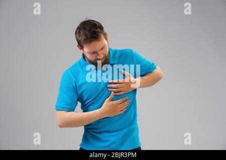 Der junge kaukasische Mann leidet unter Herzschmerz, fühlt sich krank, krank und Schwäche isoliert auf grauem Studiohintergrund. Symptome von Krankheit oder ungesunder Lebensweise. Krankheit, Unmut, traurig und verärgert. Copyspace. Stockfoto