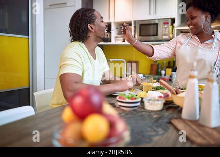 Junge Frau füttert ein Männchen in der Küche, lächelnd, eine gute Zeit beim Essen Stockfoto