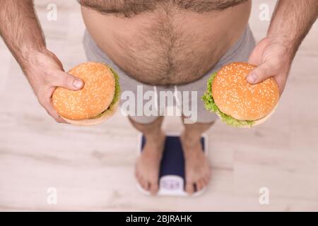 Übergewichtiger Mann hält Burger während der Messung sein Gewicht mit Waage auf dem Boden Stockfoto