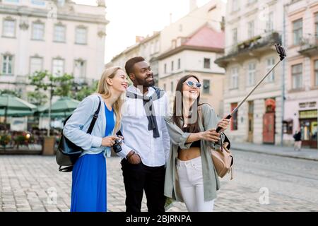 Multiethnische Freunde, Touristen, junge Afrikaner und zwei kaukasische Frauen, tragen Casual Wear erkundet neue Stadt zusammen, lächelt und macht Foto Stockfoto