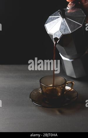 Frauenhand, die Kaffee aus der italienischen Kaffeemaschine in einer braunen Glastopf, schwarzer Hintergrund gießt. Kaffee am Morgen Stockfoto