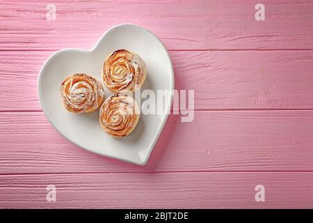 Herzförmiger Teller mit Apfelrosen aus Blätterteig auf farbigem Holzhintergrund Stockfoto