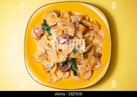 Butterfly Pasta mit gebratenen Pilzen, grüner Spinat auf einem gelben Teller auf orangefarbenem Hintergrund Stockfoto