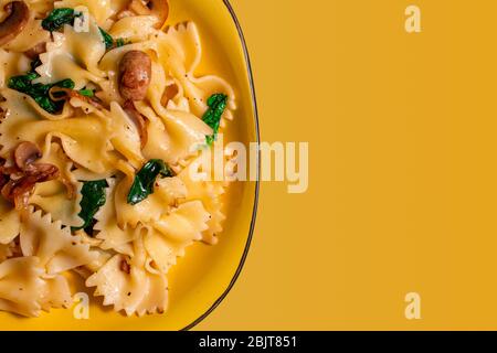 Schmetterling Pasta mit gebratenen Pilzen, grüner Spinat auf einem gelben Teller auf einem orangefarbenen Hintergrund. Copy space Stockfoto