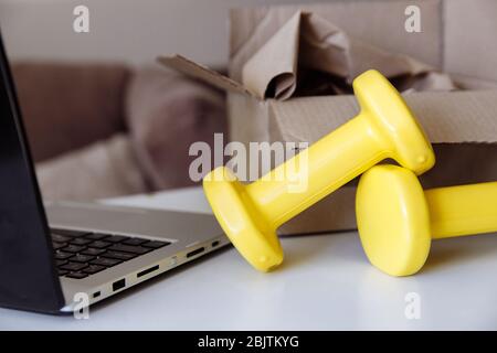 Zwei gelbe Hanteln liegen auf einem weißen Tisch. Daneben befindet sich ein Laptop. Im Hintergrund befindet sich eine Lieferbox. Stockfoto