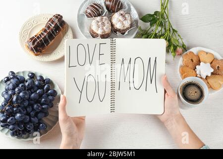 Frau hält Notizbuch mit Worten 'Love You Mama' am Tisch. Frühstück für den Muttertag Stockfoto