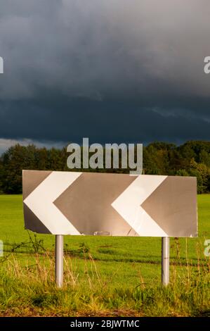 Ein Schild mit Chevron warnt vor einer scharfen Kurve auf einer Landstraße, hinter der dunkle Gewitterwolken stehen. Stockfoto