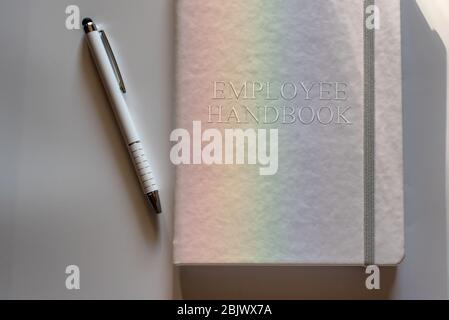 Weißes Mitarbeiterhandbuch oder Handbuch mit weißem Stift auf weißer Oberfläche, die durch ein Licht in Regenbogen beleuchtet wird - Richtlinie zur Personalverwaltung Stockfoto