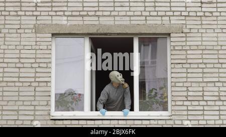 Der alte Mann bleibt am Fenster in der Gasmaske. Isoliert zu Hause auf Quarantäne. Virenausbruch. Älterer Großvater. COVID-19 Prävention gegen pandemische Coronavirus. Soziale Distanz. Konzept „Bleib zu Hause“ Stockfoto