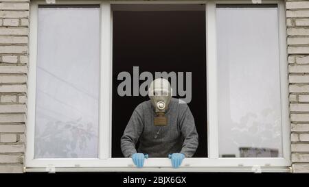 Der alte Mann bleibt am Fenster in der Gasmaske. Isoliert zu Hause auf Quarantäne. Virenausbruch. Älterer Großvater. COVID-19 Prävention gegen pandemische Coronavirus. Soziale Distanz. Konzept „Bleib zu Hause“ Stockfoto