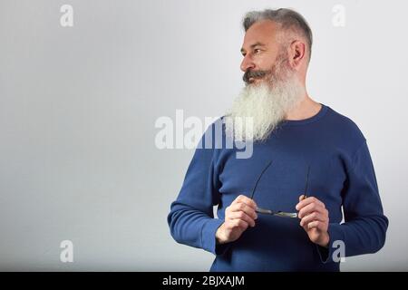 Portrait des reifen grauhaarigen bärtigen Mannes in blauem Sweatshirt mit Brille in der Hand sieht weg auf grauem Hintergrund, selektive Fokussierung Stockfoto
