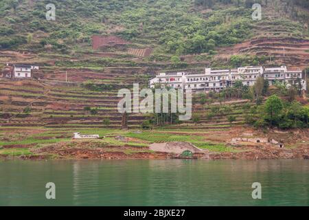 Wuchan, China - 7. Mai 2010: Dicui oder Emerald Gorge am Daning River. Weiße Häuser des Dorfes auf braunen terrassierten landwirtschaftlichen Grundstücken über grünem Wasser gesetzt Stockfoto