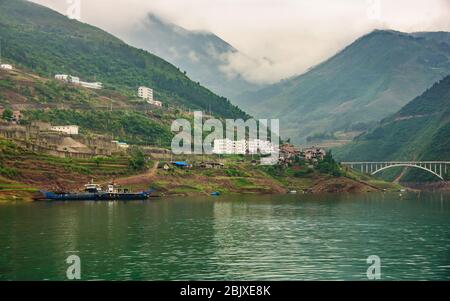 Wuchan, China - 7. Mai 2010: Dicui oder Emerald Gorge am Daning River. Landschaft mit Bergen unter Wolkenlandschaft mit alten und neuen Wohnungen, blaue Fähren Stockfoto