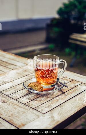 Heißer grüner Tee in Glasbecher mit Teelöffel und gebissenen Plätzchen. Auf einem alten Holztisch draußen im Hinterhof. Isolierung. Gesunde Lebensweise. Stockfoto