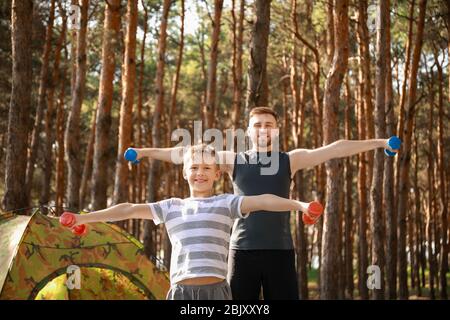 Vater und sein kleiner Sohn Training mit Hanteln in der Nähe von Camping Zelt im Wald Stockfoto