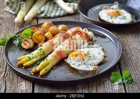 Gebackener weißer Spargel in Schinkenstreifen gewickelt, serviert mit knusprigen Kartoffeln und Spiegeleiern auf einem Holztisch Stockfoto