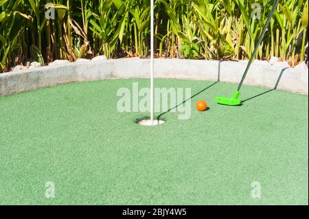 Green Mini Golfclub setzt und orange Ball in Richtung eines Lochs auf grünem Rasen Stockfoto