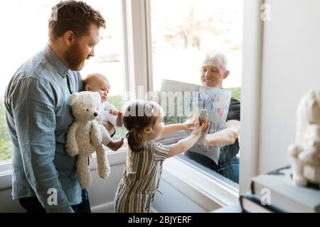 Großvater besucht Familie mit Enkeln (2-3 Monate, 2-3) und zeigt Happy Birthday Zeichen durch Fenster Stockfoto