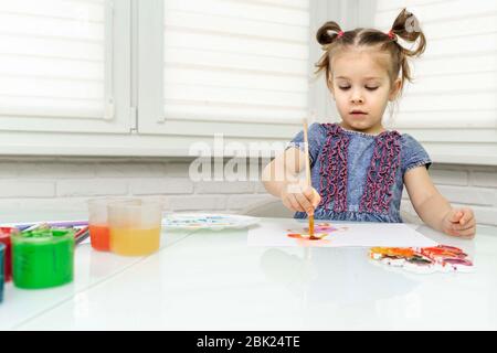Ein kleines Mädchen in einem blauen Kleid malt am Fenster, hat einen Pinsel genommen und zeichnet ein Bild für ihren Vater. Bleiben Sie zu Hause, Virus covid-19, Selbstisolation, isolat Stockfoto