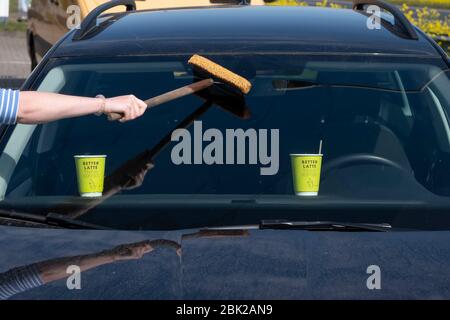 Frau reinigt Vorderglas des Autos mit einem Schwamm und Wasser. Yelllow Papierbecher im Auto mit Text. Auf dem Armaturenbrett vor der Vorderseite stehen Stockfoto