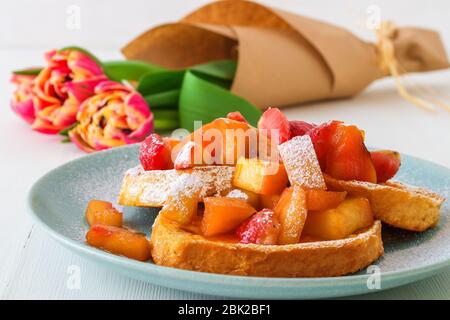 Französischer Toast mit geschmorten Äpfeln und Erdbeeren, mit einer Tasse Kaffee und Tulpenblumen auf einem weißen Holztisch. French Toast oder auch Pain perdu o Stockfoto