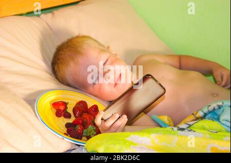 Morgen in der Hütte - ein kleiner Junge mit blonden Haaren und blauen Augen liegt auf dem Bett, glücklich vor dem Sonnenlicht zu schielen und Cartoons im ph-wert zu beobachten Stockfoto