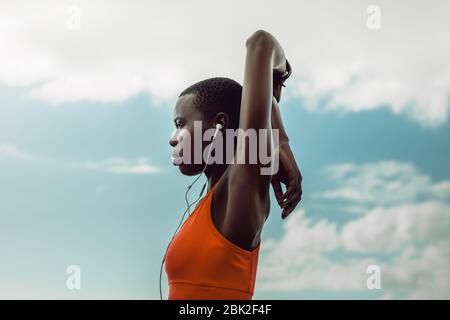 Afrikanische Frau mit Ohrhörern tun Hand Stretching Übung im Freien. Frauen in Sportbekleidung beim Warmup-Workout draußen gegen den Himmel.