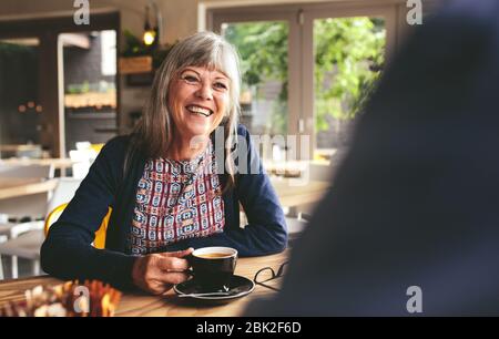 Fröhliche ältere Frau sitzt am Tisch im Café und spricht mit einem Mann, der herumsitzt. Glücklich reifen Frau im Café. Stockfoto