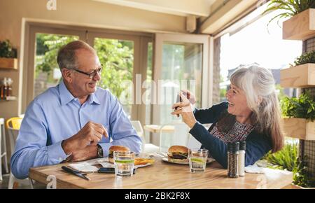 Lächelnde ältere Frau, die in einem Restaurant Fotos von ihrem Mann machte. Rentnerpaar genießen in einem Café Picutres mit einem Handy klicken. Stockfoto