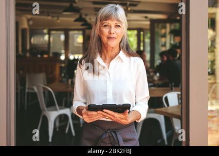 Ältere Frau, die in einem Café arbeitet, mit einem digitalen Tablet an der Tür steht. Ältere Frau, die als Kellnerin in einem Restaurant arbeitet. Stockfoto