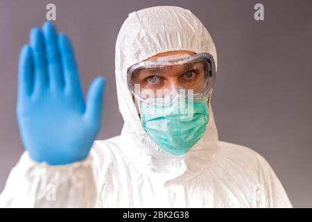 Männlicher Arzt mit chirurgischer Maske, Schutzbrille und Schutzanzug, der den Anschlag auf grauem Hintergrund zeigt. Stockfoto
