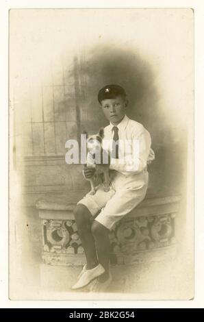 Postkarte des Jungen aus dem frühen 1900, der Mütze, Shorts, Cricket-Weiße trägt, einen Terrier in der Hand, London, Großbritannien um 1918 Stockfoto