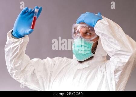 Männlicher Arzt mit Operationsmaske, Schutzbrille und Schutzanzug, der Blutproben auf grauem Hintergrund hält. Fläschchen mit Cure Covid-19. Stockfoto