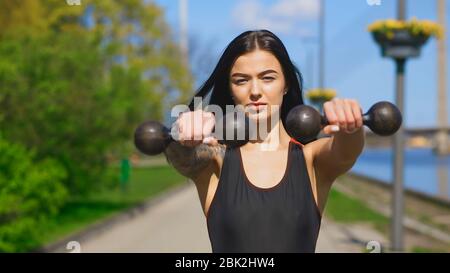 Fit Brünette Frau hält zwei alte Hanteln während sonnigen Tag und schaut Sie an Stockfoto
