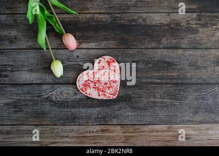 Eine herzförmige rosa Schokolade mit getrockneten Erdbeeren auf einem Vintage dunklen Holztisch Hintergrund mit Tulpenblüten. Liebe und Grußkarte Konzept. Stockfoto