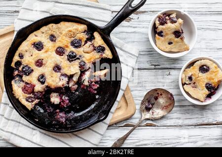 Frisch gemachter, hausgemachter Kirschpflasterpflasterer, in einer gusseisernen Pfanne mit antikem Löffel und zwei Portionen auf einem rustikalen Tisch aus weißem Holz gebacken. Bild aufgenommen von oben vi Stockfoto