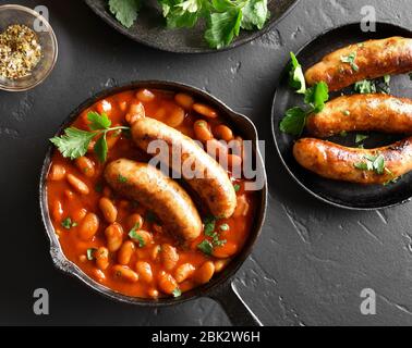 Gegrillte Würstchen mit gebackenen weißen Bohnen in Tomatensoße in der Pfanne über schwarzen Stein. Ansicht von oben, flach Stockfoto