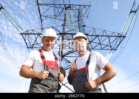 Zwei Erwachsene männliche Elektriker zeigen wie. Zwei Arbeiter von Kraftwerken auf dem Hintergrund eines elektrischen Pylons. Elektrische Unterstützung 8000 Kilowatt Stockfoto