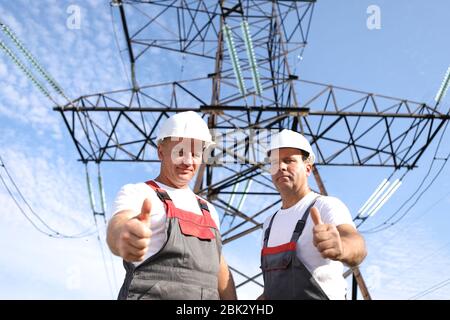 Zwei Erwachsene männliche Elektriker zeigen wie. Zwei Arbeiter von Kraftwerken auf dem Hintergrund eines elektrischen Pylons. Elektrische Unterstützung 8000 Kilowatt Stockfoto