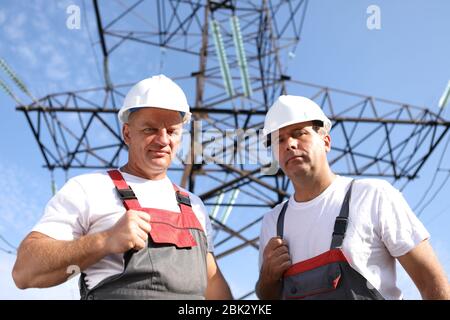 Zwei Erwachsene männliche Elektriker zeigen wie. Zwei Arbeiter von Kraftwerken auf dem Hintergrund eines elektrischen Pylons. Elektrische Unterstützung 8000 Kilowatt Stockfoto