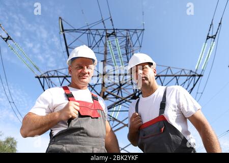 Zwei Erwachsene männliche Elektriker zeigen wie. Zwei Arbeiter von Kraftwerken auf dem Hintergrund eines elektrischen Pylons. Elektrische Unterstützung 8000 Kilowatt Stockfoto