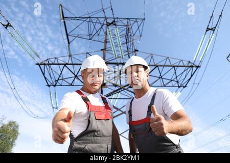 Zwei Erwachsene männliche Elektriker zeigen wie. Zwei Arbeiter von Kraftwerken auf dem Hintergrund eines elektrischen Pylons. Elektrische Unterstützung 8000 Kilowatt Stockfoto