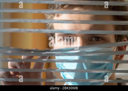 Ein junger Mann in einer medizinischen Gesichtsmaske blickt durch die Jalousien aus dem Fenster. Quarantäne während einer Epidemie. Stockfoto