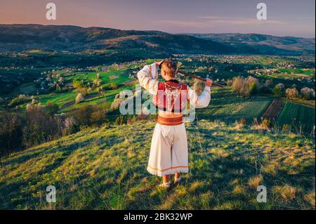 Mann in traditioneller Volkskleid, der das Licht des Sonnenaufgangs in einer wunderschönen, warmen Frühlingslandschaft betrachtet. Ländliche Wiese und Frühlingsblüten Kirschbäume im Hintergrund Stockfoto